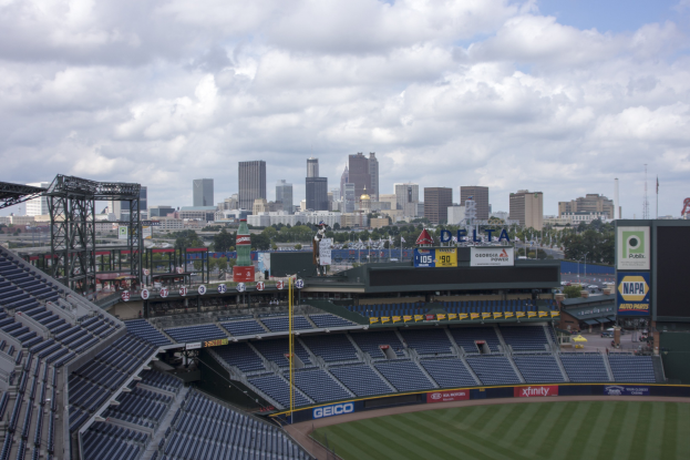 Baseballstadion mit Stadtpanorama im Hintergrund, gefüllt mit Stühlen, Pfosten und Brettern, auf Gras mit Bäumen und Gebäuden in der Ferne unter einem bewölkten Himmel.