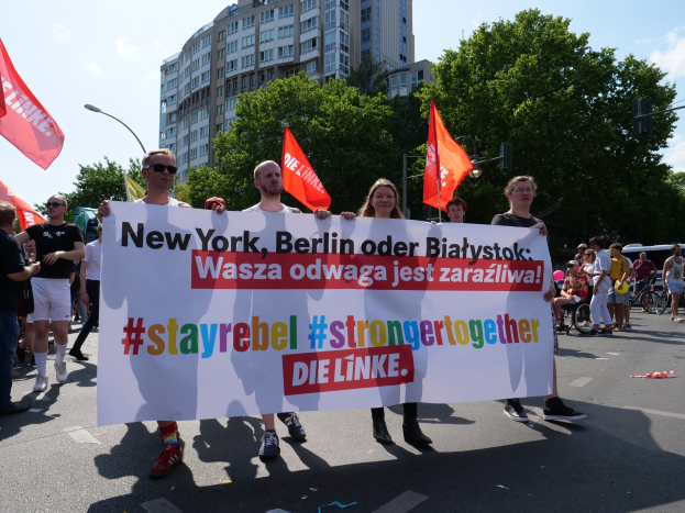 Gruppe von Menschen, die eine Straße in Berlin, Deutschland, entlanggehen und ein Banner mit der Aufschrift "Stay Rebel Stronger Together" tragen, mit Bäumen, Laternenpfählen und Gebäuden, die die Straße säumen, unter einem bewölkten Himmel.