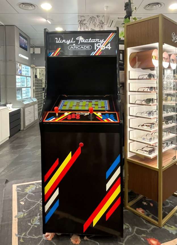 Ein Geschäft mit einem zentralen Retro-Arcade-Automaten, einem rechten Glasvitrinenschrank und Hintergrundschränken, Fernseher und anderen Gegenständen sowie Deckenleuchten, die eine Retro-Atmosphäre schaffen.