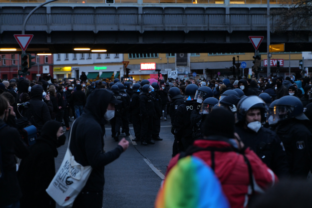 Eine große Gruppe von Menschen vor einer Reihe von Polizisten, einige mit Helmen und Taschen oder Kameras, mit Gebäuden, Schildern, Laternen, Verkehrsampeln, einer Brücke und einem Baum im Hintergrund.