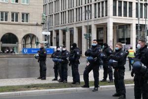 Eine Gruppe von Polizisten in schwarzen Uniformen und Masken steht vor einem Berliner Gebäude mit Glasfenstern und Säulen, einige halten Helme, mit Laternenmasten, Verkehrsampeln, Texttafeln, einer Statue und anderen Menschen im Hintergrund auf einem grasbewachsenen Boden.