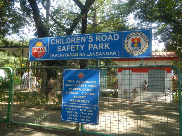 Kindersicherheitspark mit Metallzaun, zwei blauen Informationsschildern, einem kleinen Haus, Pflanzen, Bäumen und einem bewölkten Himmel.