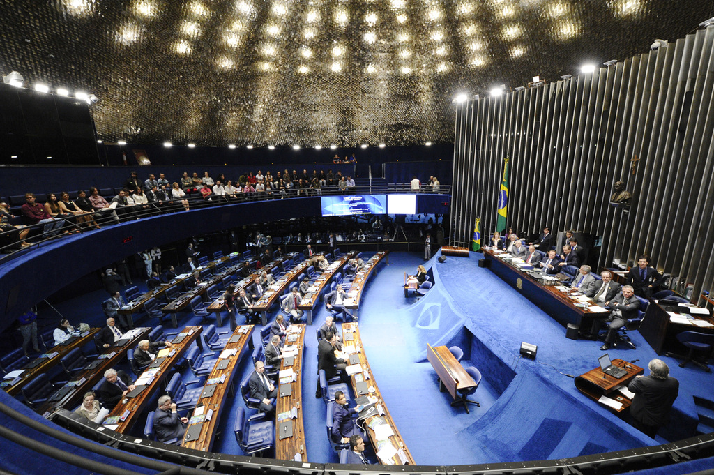 Eine Konferenzbühne mit sitzenden Personen an einem Tisch mit Papieren und Gegenständen, flankiert von Fahnen und einer Skulptur im Hintergrund, mit Zuschauern an Tischen und hinter einer Absperrung mit Bildschirmen, unter dekorativen Deckenleuchten.
