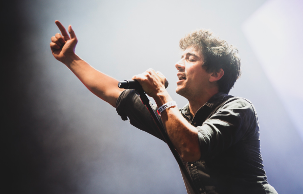 Ein Mann in schwarzem Hemd und Jeans singt in ein Mikrofon auf einer hell erleuchteten Bühne, seine Augen geschlossen, während das Publikum im Hintergrund jubelt und klatscht.