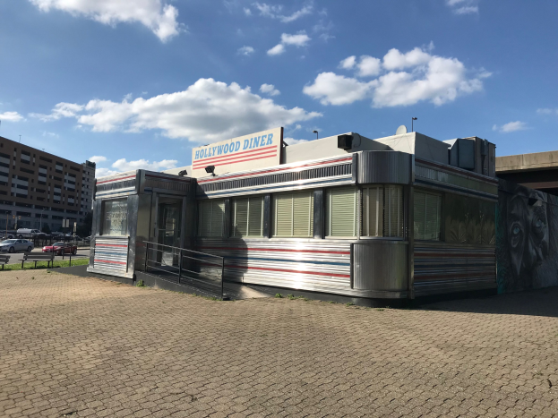 Außensicht des Hollywood Diners mit seiner Leuchtschrift, umgeben von Gebäuden, Straßenmöbeln, Fahrzeugen und einem bewölkten Himmel.