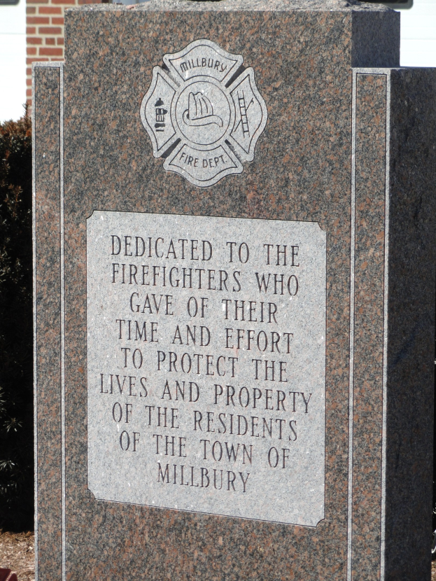 Denkmalstein mit einer Gedenktafel vor einem Backsteingebäude, umgeben von Pflanzen und Bäumen.