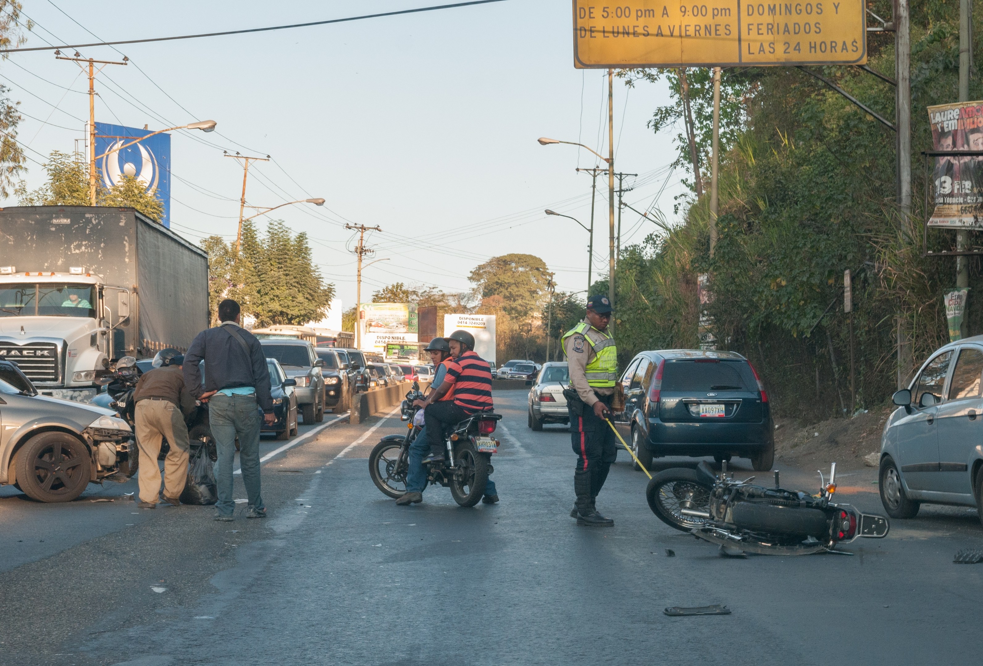 Eine Gruppe von Menschen steht in der N├Ąhe eines verunglückten Motorrads am Stra├čenrand mit mehreren Fahrzeugen, darunter ein Lastwagen, im Hintergrund und B├Ąumen, Masten, Lichtern, Schildern und Himmel.