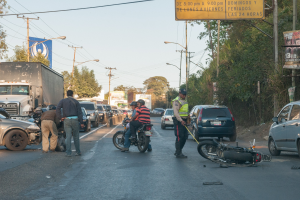 Eine Gruppe von Menschen steht in der N├Ąhe eines verunglückten Motorrads am Stra├čenrand mit mehreren Fahrzeugen, darunter ein Lastwagen, im Hintergrund und B├Ąumen, Masten, Lichtern, Schildern und Himmel.
