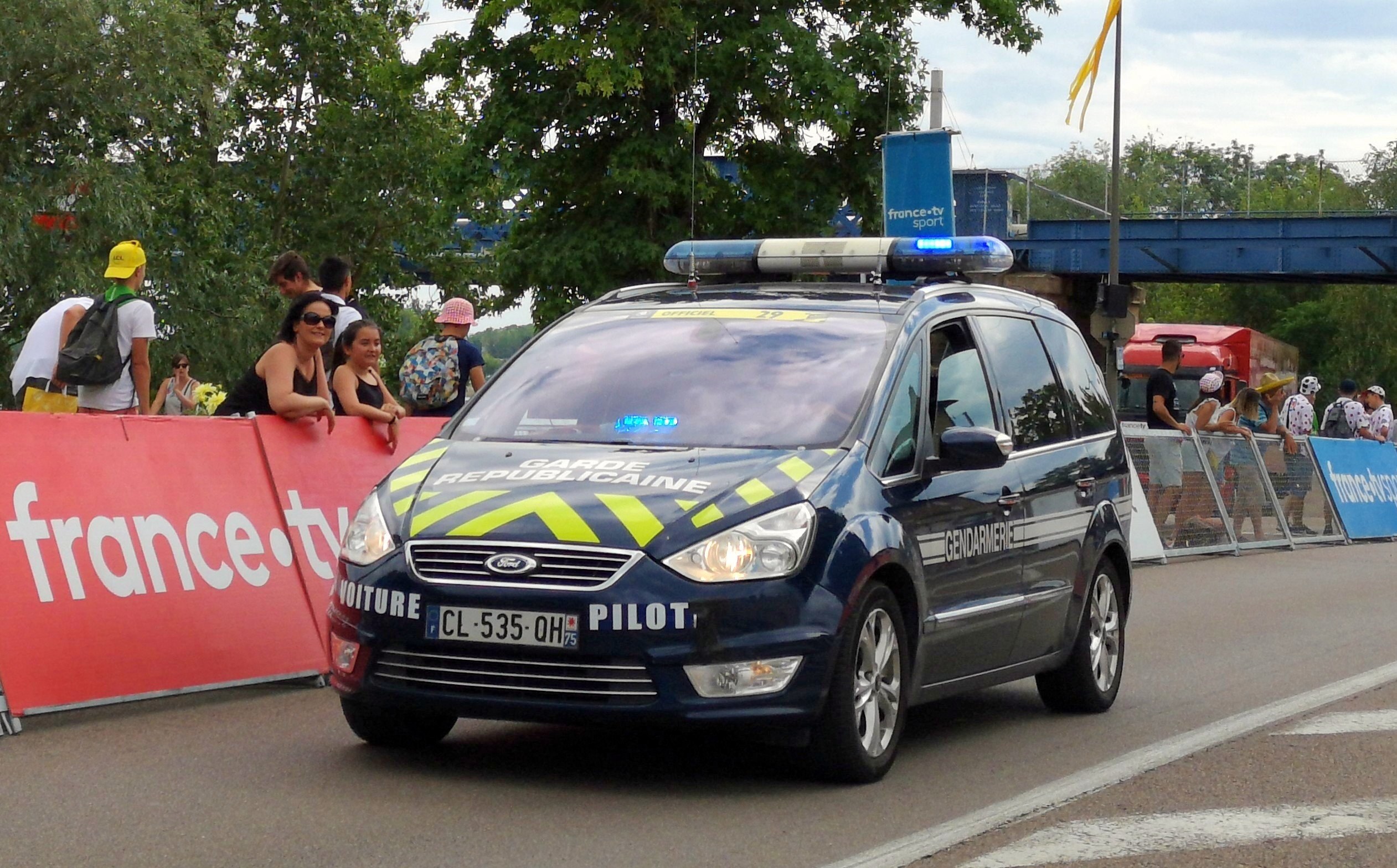 Ein Polizeiauto fährt an einer Menge vorbei, die Schilder mit Texten halten, mit Bäumen, einer Brücke, einer Flagge und Wolken im Hintergrund.