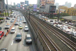 Eine belebte Straße mit Schwerlastverkehr neben einer Bahnschiene, mit Geb"uden, Werbetafeln, Masten, Lichtern, B"umen, Pflanzen, Gras und einem sichtbaren Auto und Rad im Vordergrund.