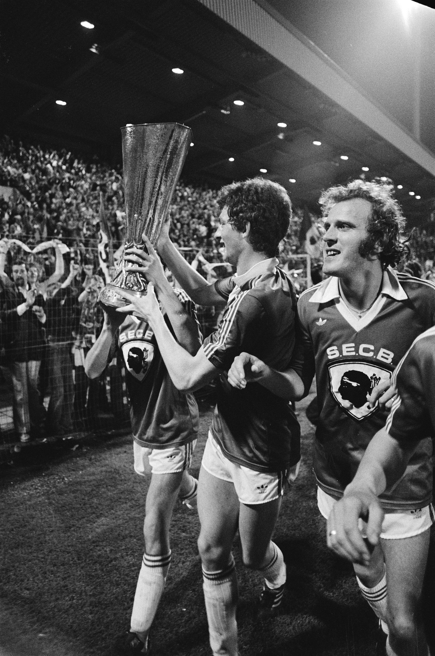 Schwarzes Foto von Fußballspielern, die mit einem Pokal auf einem Feld feiern, umgeben von einer Menge hinter einem Zaun unter Stadionlichtern.