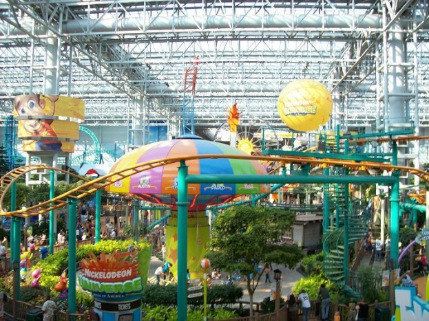 Ein großes Indoor-Wasserpark mit einer Achterbahn, umgeben von Menschen, Bäumen, Pflanzen und beleuchteten Deckenstangen und -lichtern.
