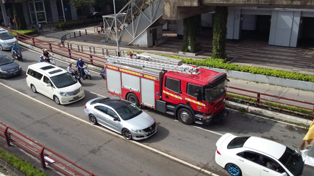 Feuerwehrfahrzeug fährt auf einer Straße mit Autos, Motorrädern und anderen Fahrzeugen; Gebäude und Bäume im Hintergrund.