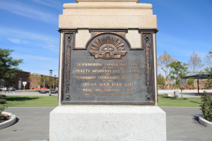 Ein Denkmal in einem Park mit einer Tafel, die 'Treue, Hingabe und Opfer' trägt, umgeben von Pflanzen, Bäumen, Gebäuden, Pfählen, Fahrzeugen, unter einem klaren blauen Himmel.