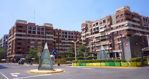 Eine Stadtstraße mit einem zentralen Denkmal, hohen Gebäuden, Straßenmöblierung, Grünflächen, Passanten und einem klaren blauen Himmel.