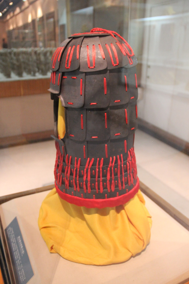 Helm, der unter Glas mit einem gelben Tuch darüber ausgestellt wird, begleitet von einer Texttafel links und Deckenlampen im Hintergrund.