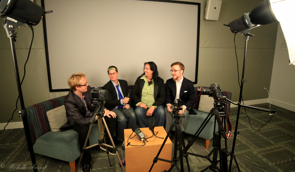 Eine Gruppe von drei Personen sitzt auf einem Sofa und gibt ein Interview. Links sitzt eine Person in einem schwarzen Mantel und Hose, in der Mitte eine Person in einem Mantel und blauer Krawatte mit einem Mikrofon und rechts eine Person, die strahlt. Auf dem Tisch in der Mitte steht ein Mikrofonrekorder, Kameras sind auf der rechten und linken Seite und große Scheinwerfer sind vorhanden, mit einem Projektionsschirm im Hintergrund.