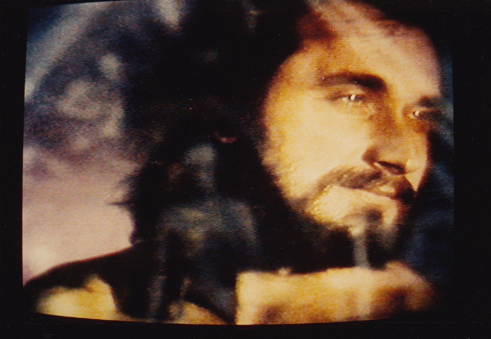 Ein Mann mit langem dunklem Bart und Schnurrbart, der ein weißes Hemd trägt, schaut ernst in die Kamera vor einem dunklen Hintergrund.