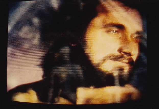 Ein Mann mit langem dunklem Bart und Schnurrbart, der ein weißes Hemd trägt, schaut ernst in die Kamera vor einem dunklen Hintergrund.