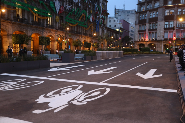 Stadtstraße mit einem Fahrradweg, umgeben von Gebäuden, Straßenmöbeln, Fußgängern auf dem Gehweg, Grünflächen und einem klaren blauen Himmel.