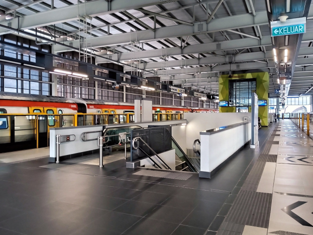 Ein Bahnhofsgebäude mit einem Zug auf den Gleisen, einem Bahnsteig mit Geländern und Treppen mit Handläufen, Informationsanzeigen, Deckenleuchten und Glaswänden.