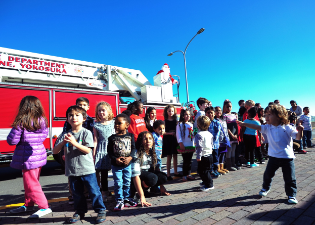 Eine Gruppe von Kindern vor einem Löschfahrzeug auf einer Straße mit Laternenpfählen, Bäumen und einem klaren blauen Himmel, das Fahrzeug wahrscheinlich Teil der Weihnachtsparade der Yokosuka-Feuerwehr.