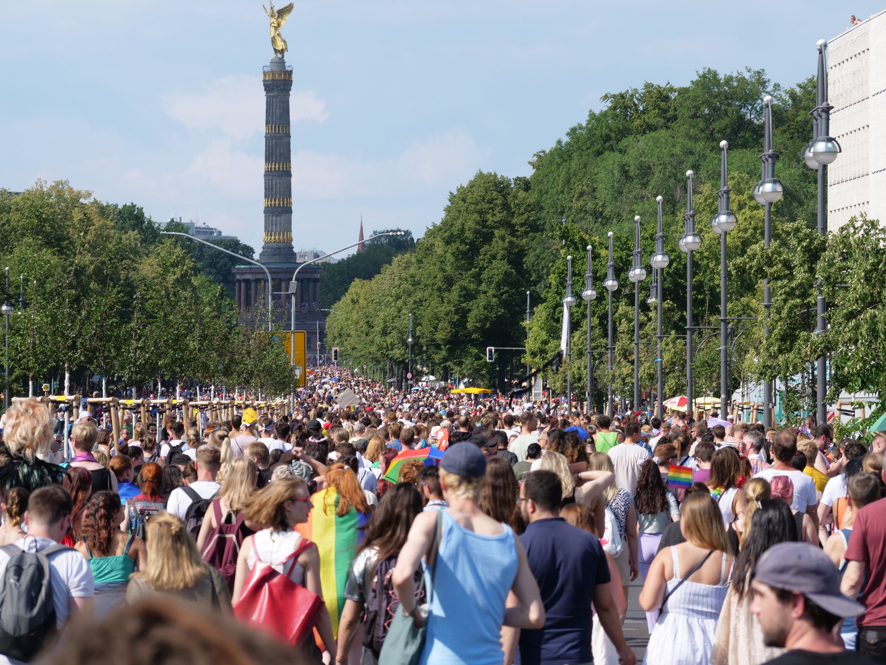 Eine große Menschenmenge geht eine Straße in Berlin, Deutschland, entlang, mit Laternenpfählen an der Straße, Bäumen und einem Turm mit einer Statue im Hintergrund und Gebäuden unter einem bewölkten Himmel.