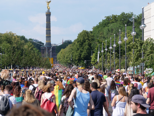 Eine große Menschenmenge geht eine Straße in Berlin, Deutschland, entlang, mit Laternenpfählen an der Straße, Bäumen und einem Turm mit einer Statue im Hintergrund und Gebäuden unter einem bewölkten Himmel.