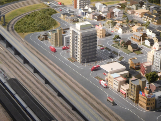 Ein Modell einer Stadtlandschaft mit einem Zug auf Schienen, umgeben von Gebäuden, Bäumen, Straßen, Fahrzeugen, Masten, Gras und einem prominenten Wolkenkratzer.