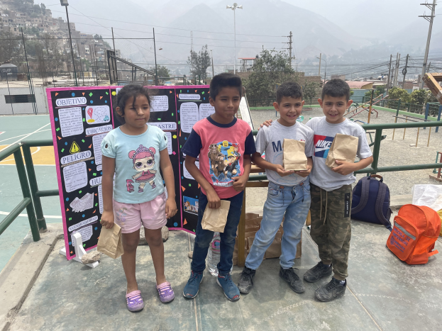 Gruppe von Kindern auf einem Basketballplatz mit Papierbeuteln in den Händen, mit Beuteln und Gegenständen rechts und einer Tafel mit Text links, vor dem Hintergrund von Zäunen, Pfählen, Straßenlaternen, Strommasten, Bäumen, Gebäuden, Hügeln und Himmel.