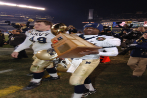 Ein Fußballspieler mit einem Pokal auf einem Feld, umgeben von einer Menge in Sportkleidung, mit Schildern, Lichtern und einem klaren blauen Himmel im Hintergrund.