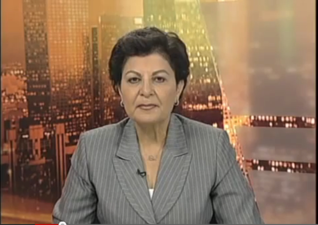 Eine Frau in einem Business-Outfit sitzt vor einem Fernseher, im Hintergrund sind ein paar Gebäude zu sehen, die als Nachrichtensprecherin mit Text am unteren Bildschirmrand erscheint.