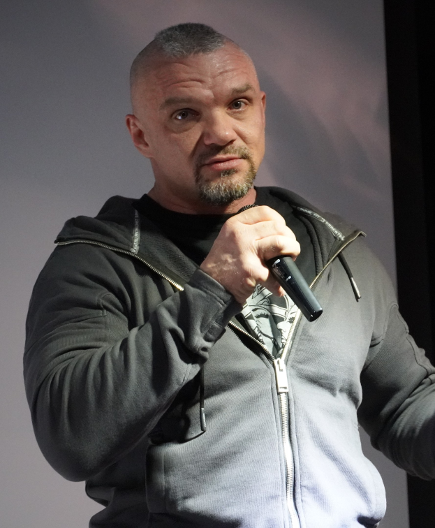 Ein Mann in einer schwarzen Kapuzenjacke und einem schwarzen Anzug mit einem wei├čen Hemd, glattem Haar und einem selbstbewussten Ausdruck halt einen Mikrofon, whrend er vor einer Wand steht.