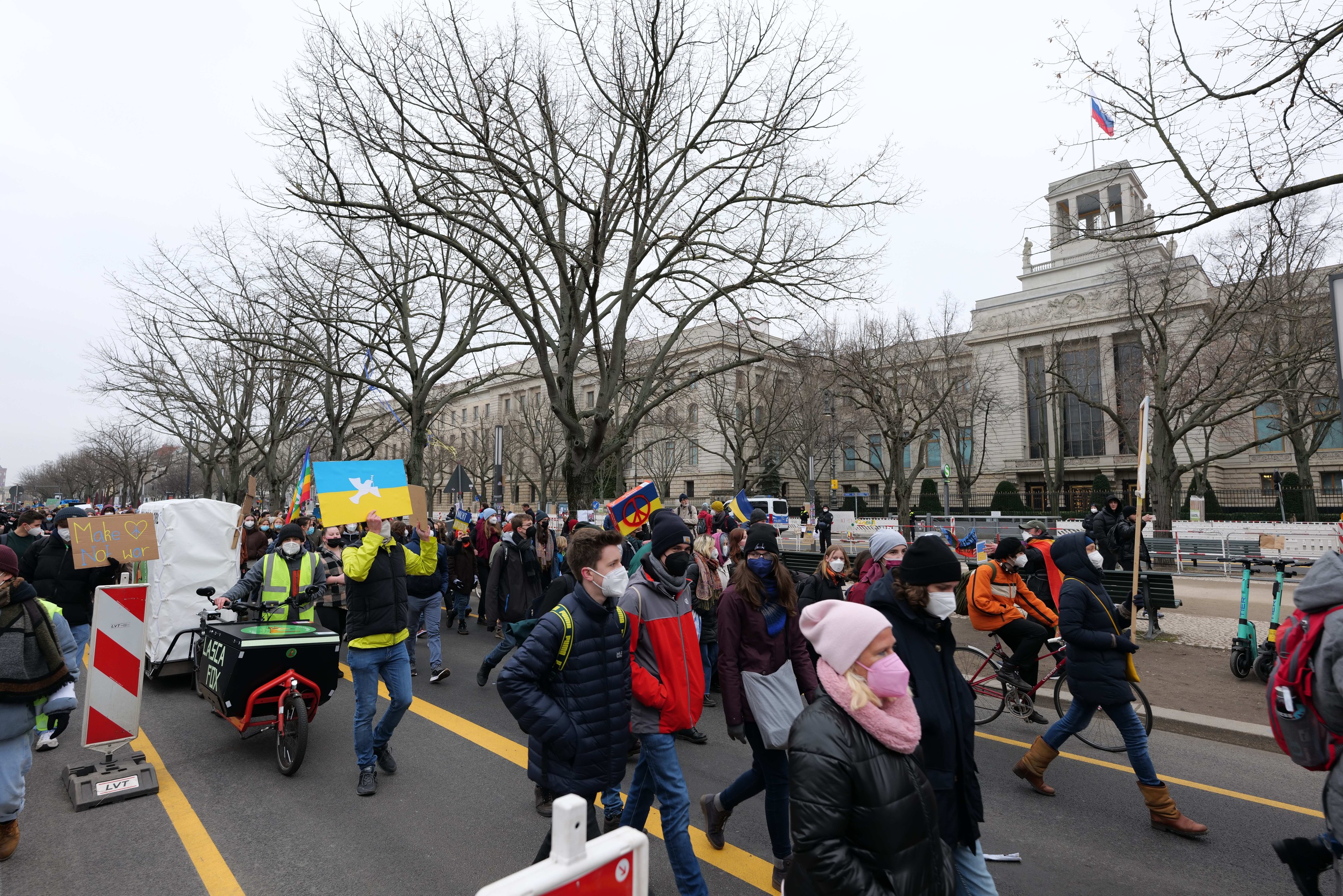 Eine große Gruppe von Menschen nimmt an einer Protestdemo in Washington, D.C. am 21.01.2020 teil und marschiert eine Straße entlang, wobei sie Schilder und Banner schwingt und einige Fahrräder fahren. Im Hintergrund sind Bäume und Schilder zu sehen, unter einem klaren blauen Himmel.
