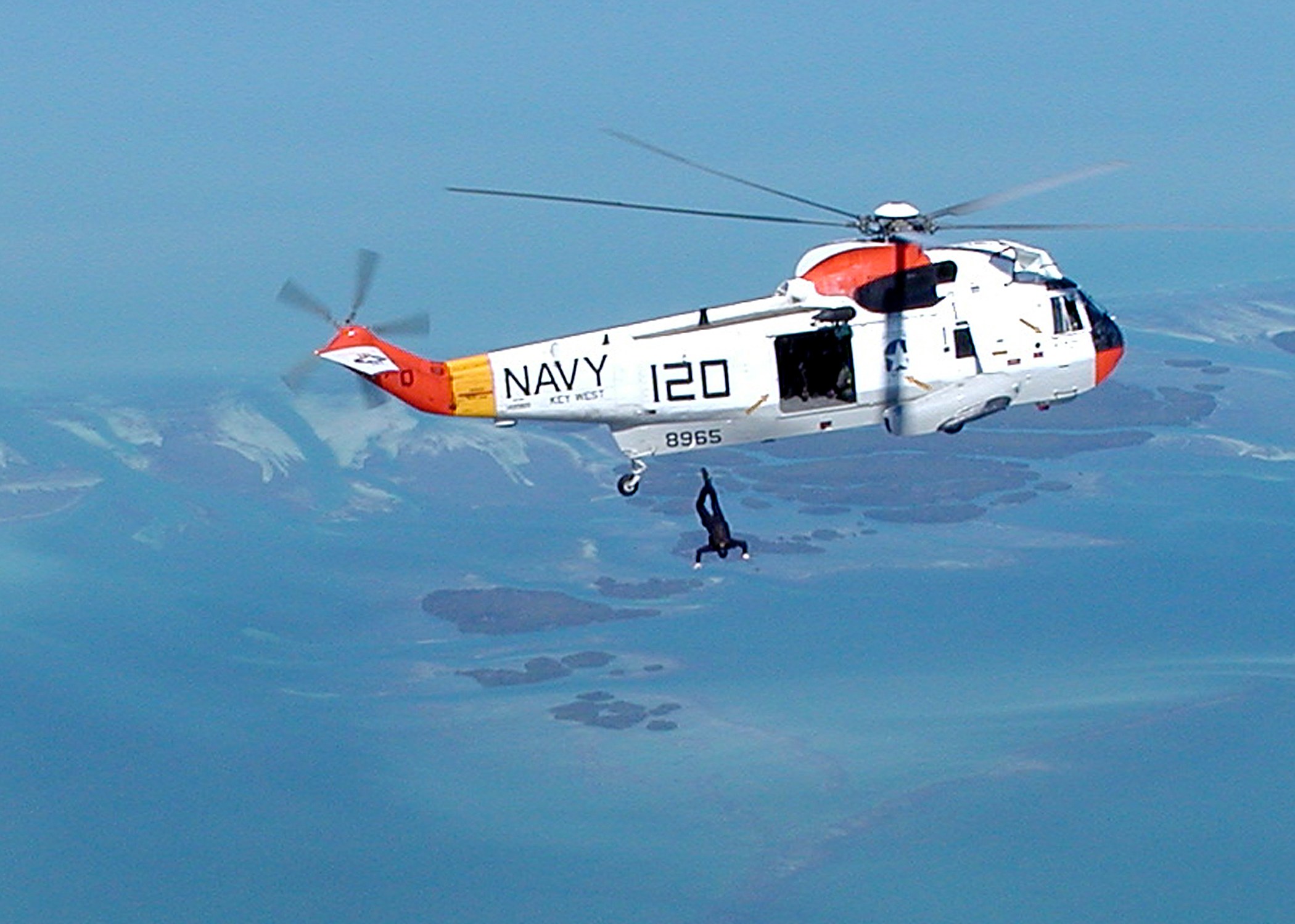 Hubschrauber fliegt über Wasser mit einer Person darunter, Himmel im Hintergrund sichtbar; Hubschrauber hat Rettungsaufschrift.