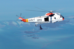 Hubschrauber fliegt über Wasser mit einer Person darunter, Himmel im Hintergrund sichtbar; Hubschrauber hat Rettungsaufschrift.