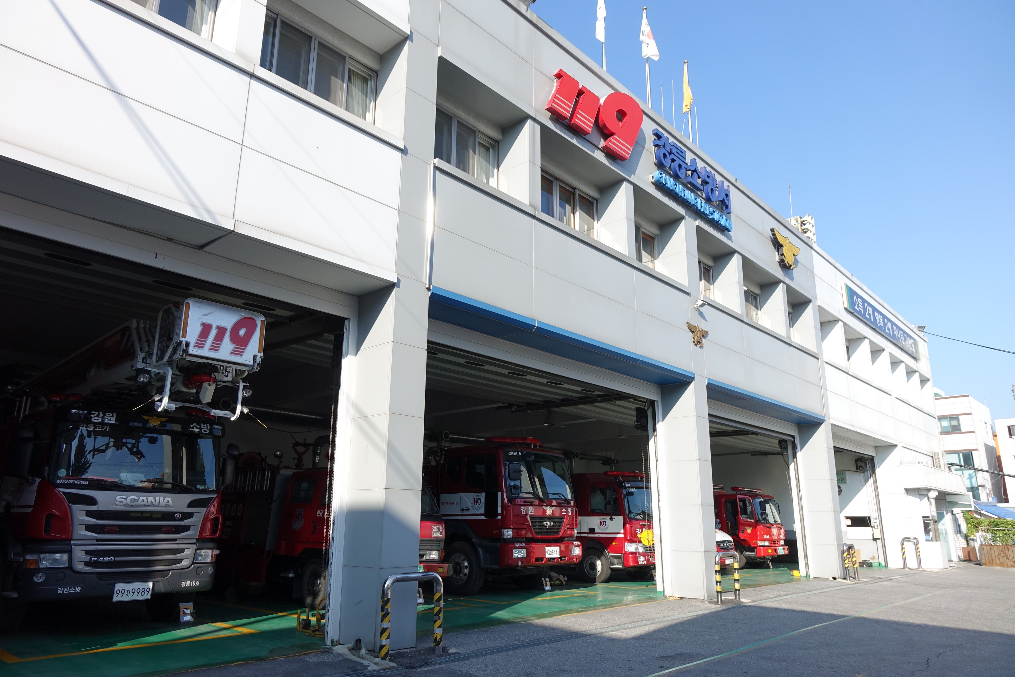 Feuerwache mit mehreren Feuerwehrautos vorne, mit Fenstern, Säulen und Namensschildern, sowie Fahnen im Vordergrund und anderen Gebäuden gegen einen klaren blauen Himmel.
