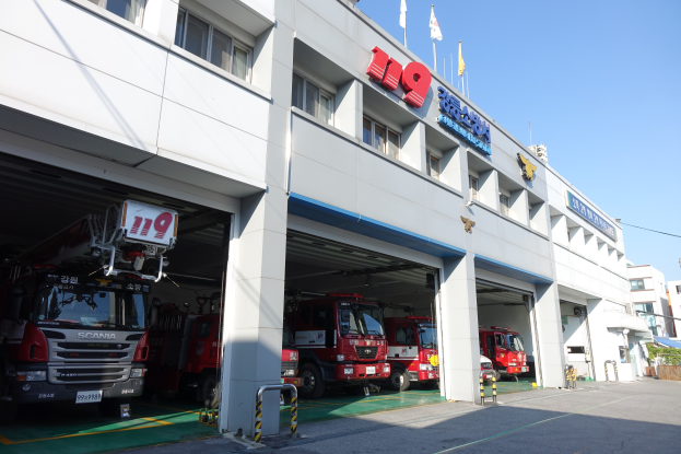 Feuerwache mit mehreren Feuerwehrautos vorne, mit Fenstern, Säulen und Namensschildern, sowie Fahnen im Vordergrund und anderen Gebäuden gegen einen klaren blauen Himmel.