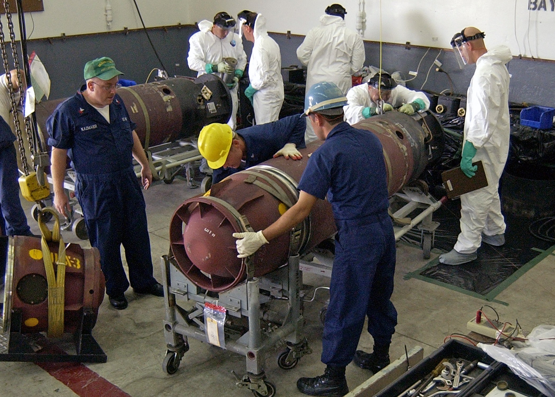 Männer in Helmen und Handschuhen, die eine große Bombe in einer Fabrik mit Maschinen und Wandtext auseinandernhmen.