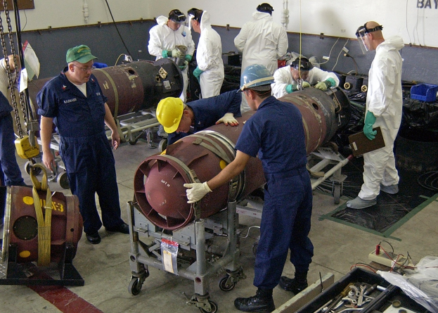 Männer in Helmen und Handschuhen, die eine große Bombe in einer Fabrik mit Maschinen und Wandtext auseinandernhmen.