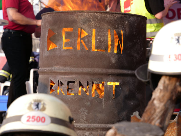 Eine Gruppe von Menschen steht um einen Feuerhydranten mit der Aufschrift "Berlin", im Vordergrund ein brennender Eimer, Helme unten und ein hölzernes Objekt rechts.