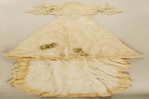 Ein weißes Brautkleid mit Rüschen, Spitzenverzierung und floraler Stickerei auf einer weißen Oberfläche, mit einem eng anliegenden Oberteil, Herzausschnitt, voluminösem Rock und langer Schleppe.