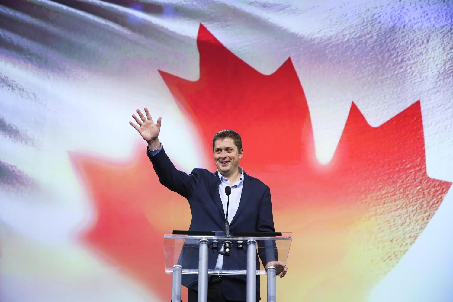 Der kanadische Premierminister Alex Salmond steht an einem Podium mit Mikrofonen, lächelt vor einer kanadischen Flagge, wobei ein Bildschirm im Hintergrund sichtbar ist.