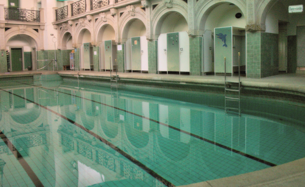 Indoor-Schwimmbad mit klarem Wasser, umgeben von gefliestem Boden, Bögen, Säulen, Geländern, Türen und Texttafeln in einem großen Gebäude.