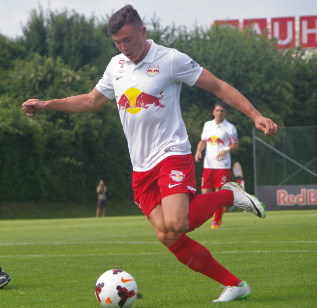 Ein professioneller Fußballspieler schießt einen Ball auf einem Rasenfeld mit Bäumen und einem klaren blauen Himmel im Hintergrund, in der Nähe einer Tafel mit der Aufschrift "RB Leipzig".