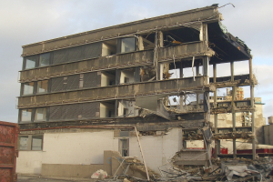 Ein teilweise abgerissenes Gebäude inmitten einer Straße, mit einem Container auf der linken Seite, zerbrochenen Fenstern und herumliegendem Schutt, unter einem bewölkten Himmel.
