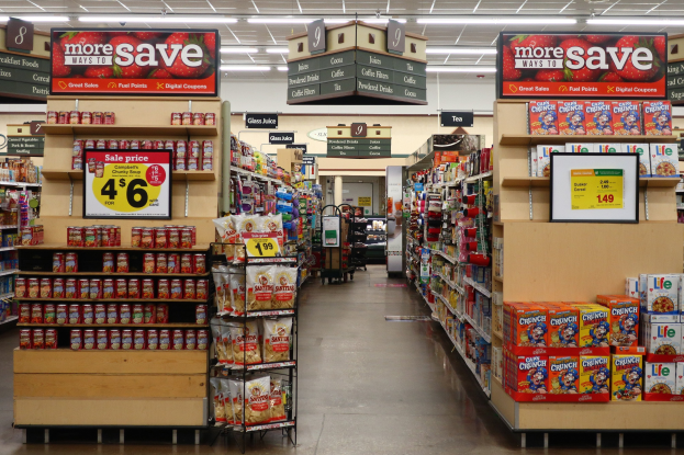 Ein Innenraum eines Lebensmittelladens mit Regalen und Boden voller verschiedener Lebensmittel, Deckenbeleuchtung und Informationsschilder im Hintergrund.