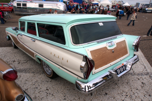 Ein gr├╝ner und wei├čer Kombi in einem belebten Parkplatz mit Zelten, Masten, B├Ąumen und einem klaren blauen Himmel im Hintergrund.