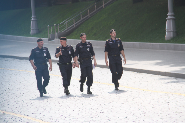 Drei Polizisten in Uniform und Mützen auf einer Straße mit Pfählen, Geländern, Treppen und Gras im Hintergrund.