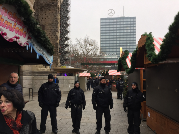 Polizeibeamte in schwarzen Uniformen stehen vor einem geschmückten Weihnachtsmarkt in Berlin, mit festlichen Deko, Gebäuden, Bäumen und einem klaren blauen Himmel im Hintergrund; ein Schild mit Text ist links sichtbar.